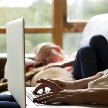 Woman using a laptop with her daughter and pet dog sleeping together next to her