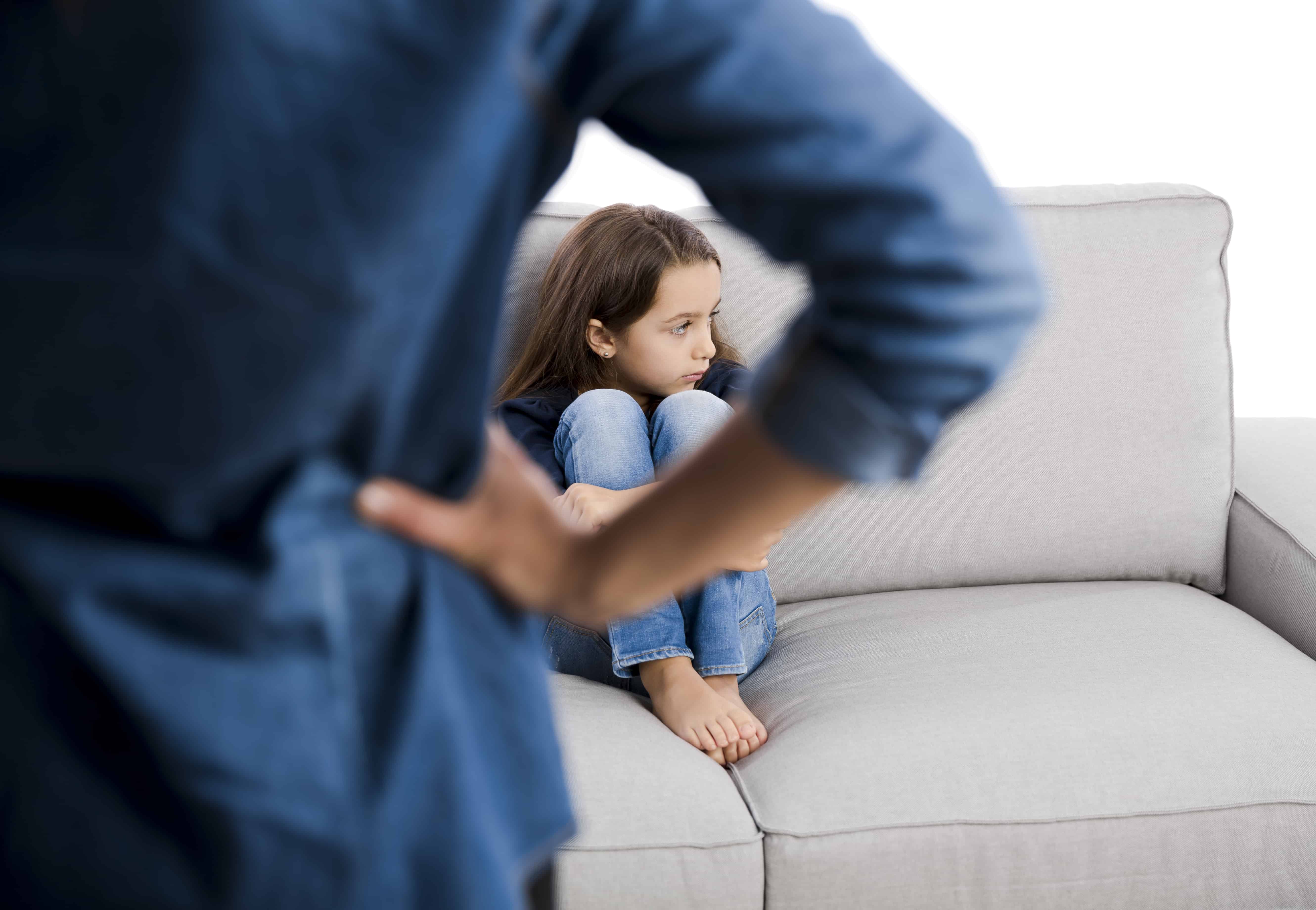 Mom with hand on hip facing upset daughter sitting on a couch.