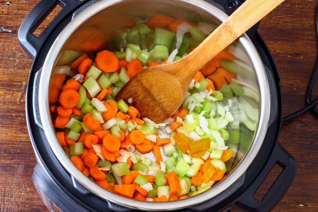 Looking into an Instant Pot with diced carrots, celery, and oinion and a wood spoon for stirring.