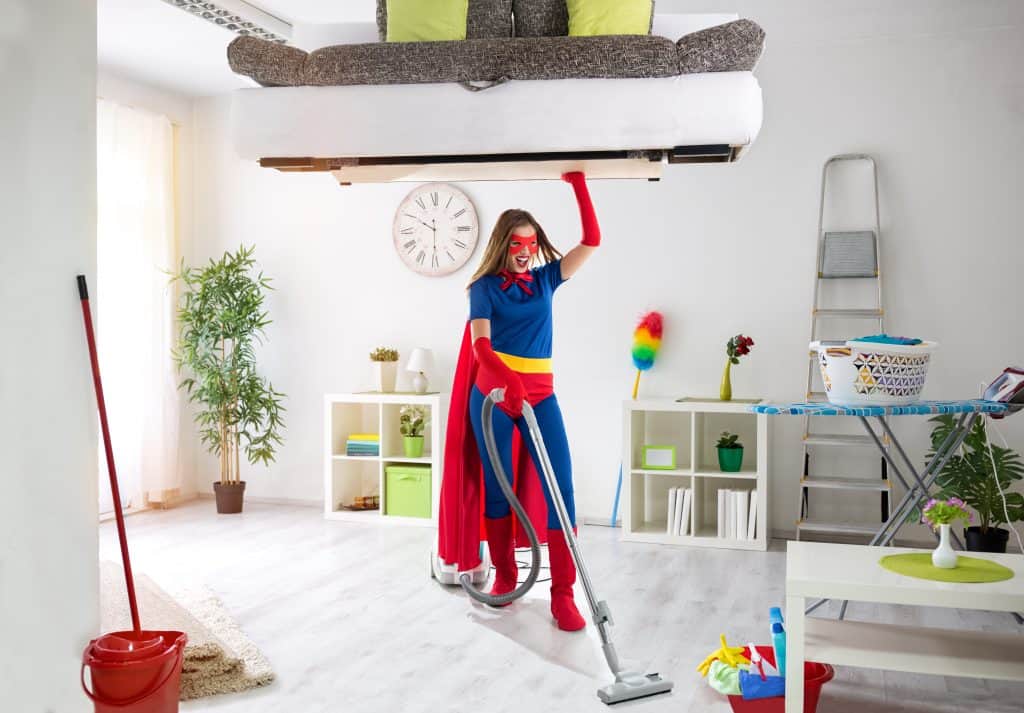 Woman dressed in super-man like outfit holding a couch above her head while vacuuming