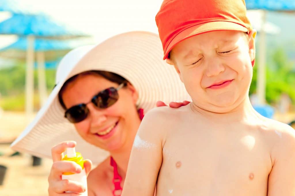 Young boy without shirt on squniting his eyes as mom sprays on sunscreen