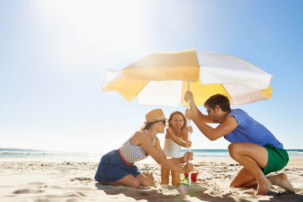 Family of 3 setting up an umbrella on the beach to shade them from the son.