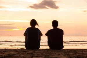 husband and wife sitting on a beach at sunset with their back to you