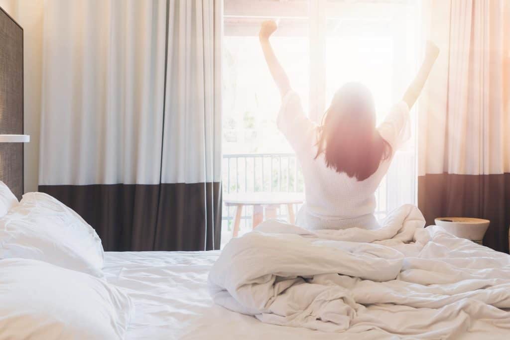 Mom waking up to start the day, sitting on a bed, looking out the window, stretching her arms.