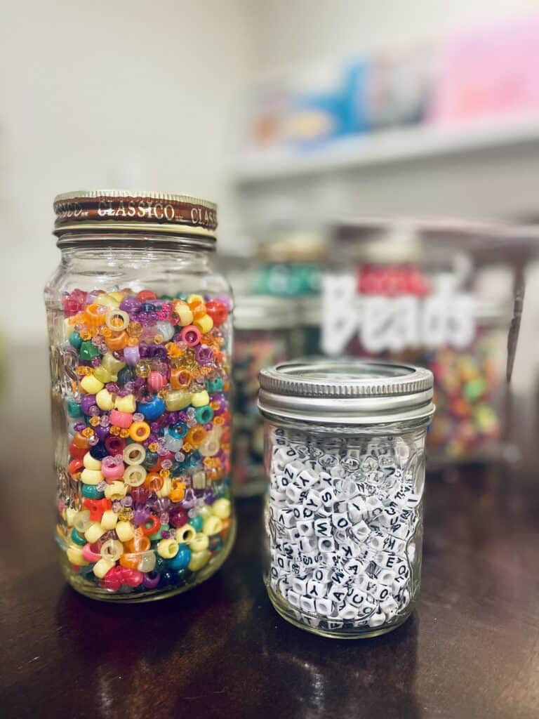 Upclose picture of an old spaghetti jar with colorful beads and an old mason jar with white beads.  Highlighting the opportunity to reuse mason jars for home organization.