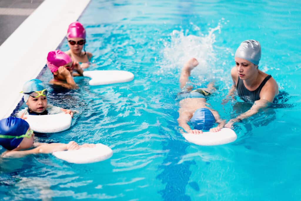 Children in swimming class, practicing with kicking boards, doing rotations, instructor helping one of them. great idea for a clutter free christmas gift.