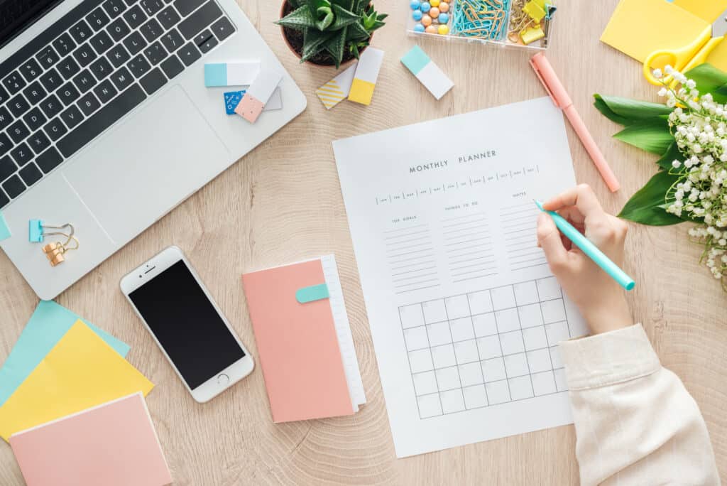 a desktop with a smartphone, a planner, and a woman's hand writing on a planner sheet