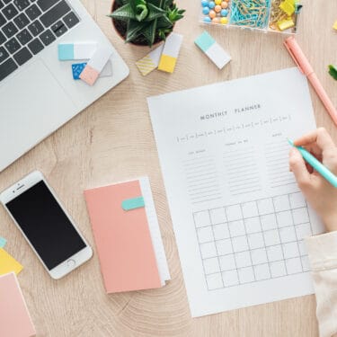 a desktop with a smartphone, a planner, and a woman's hand writing on a planner sheet
