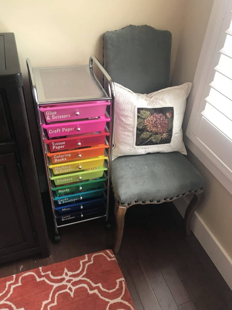 Vertical rainbow colored craft organizing cart with labels on each drawer nested in between a cabinet and a chair.