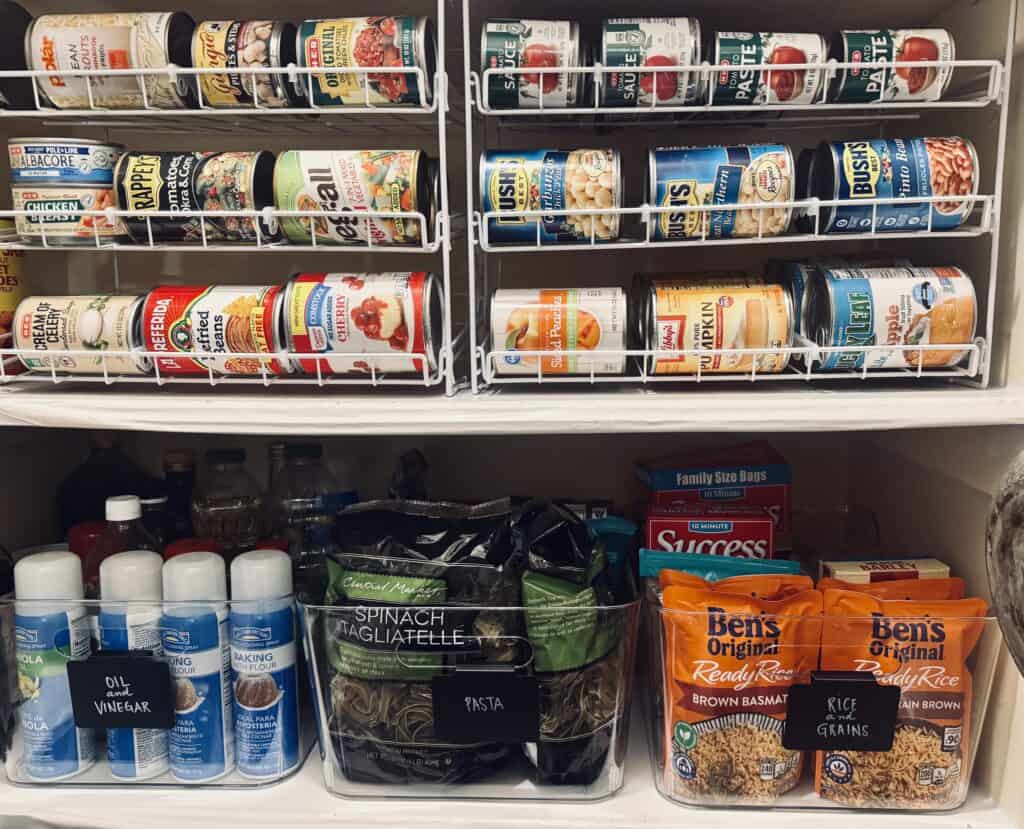 An organized deep pantry showing two shelves.  The top shelf shows wire storage housing canned goods, the second shelf shows plastic pins used for organizing oil and vinegar, pasta, and rice and grains.