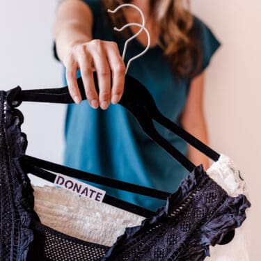 Woman holding 2 hangers with tops on them. One top has a sticker that says donate after she's asked herself 7 questions to help her decide what to keep and what to get rid of when decluttering.