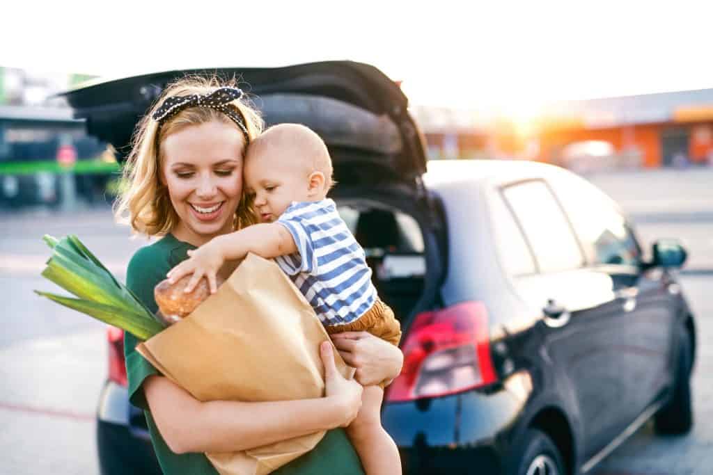 Beautiful young mother with her little baby son in front of a supermarket, holding paper shopping bag. 